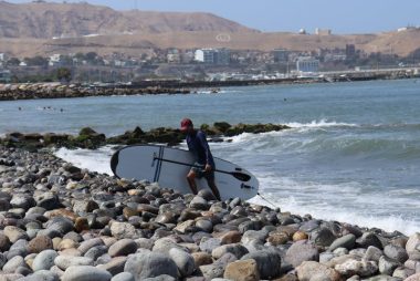 A surfer carrying a board along the rocky beach of Lima, Peru on a sunny day.