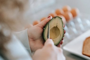 Crop anonymous female with knife cutting ripe avocado while preparing toasts at table with eggs and bread on blurred background